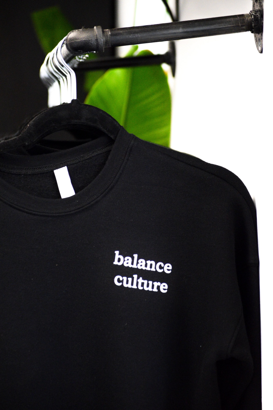 Black 'balance culture' sweatshirt hanging on a pipe rack beside a green plant