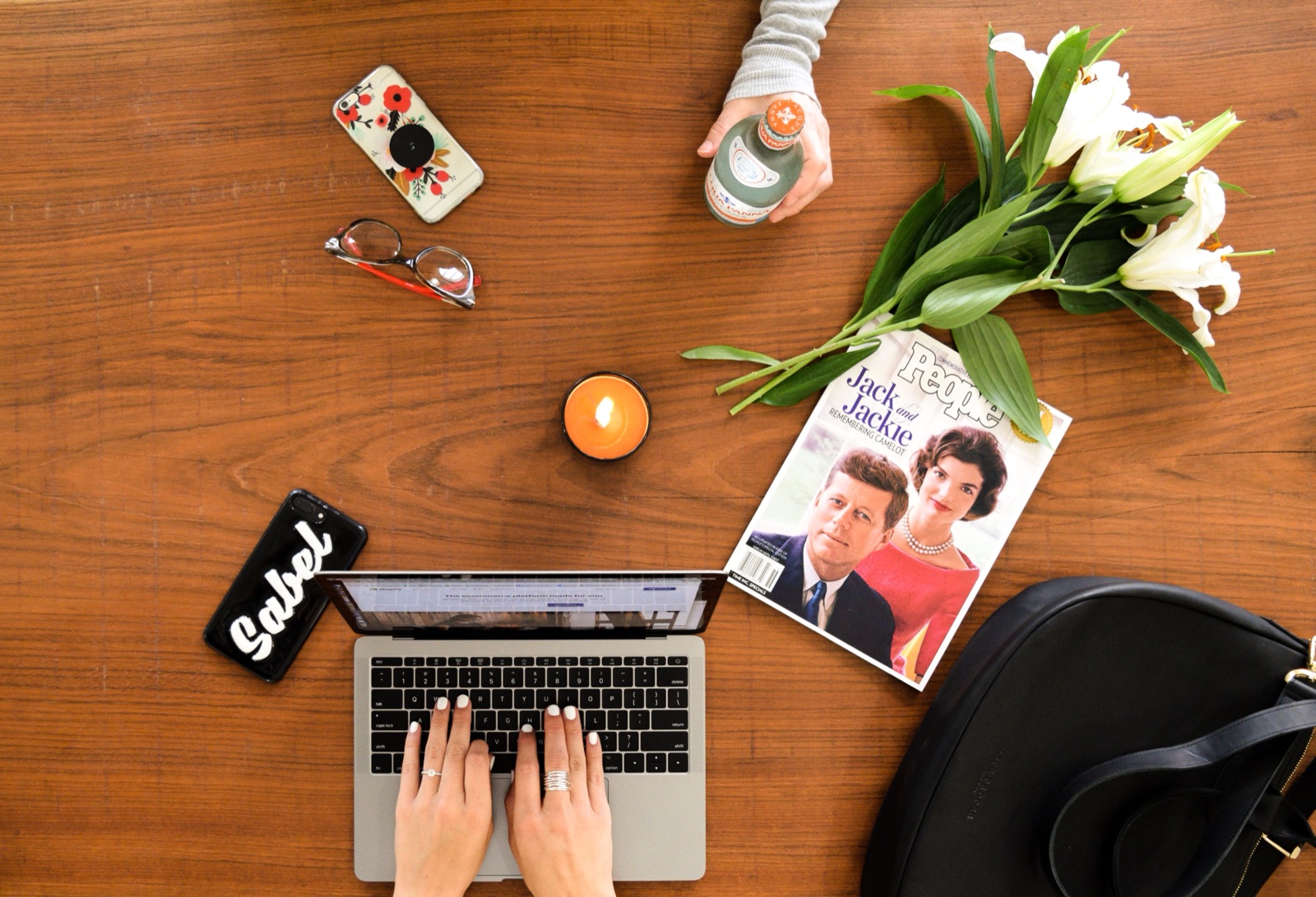 Overhead desk flatlay with a laptop, magazine, lilies, and hands typing