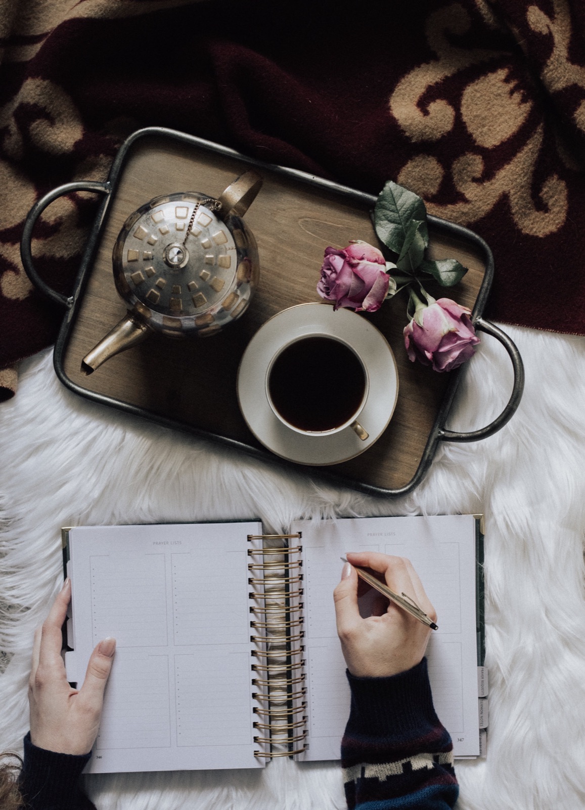 Overhead of a tea tray with roses and hands writing in a planner on a fur throw